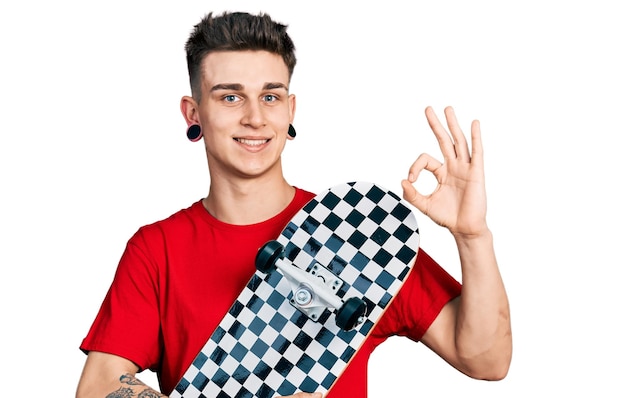 Photo gratuite jeune garçon caucasien avec dilatation des oreilles tenant skate faisant signe ok avec les doigts, souriant amical gesticulant excellent symbole
