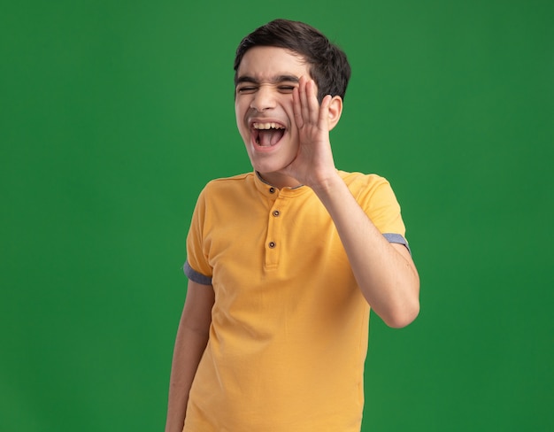 Jeune garçon caucasien gardant la main près de la bouche appelant à haute voix quelqu'un avec les yeux fermés isolé sur un mur vert avec espace de copie