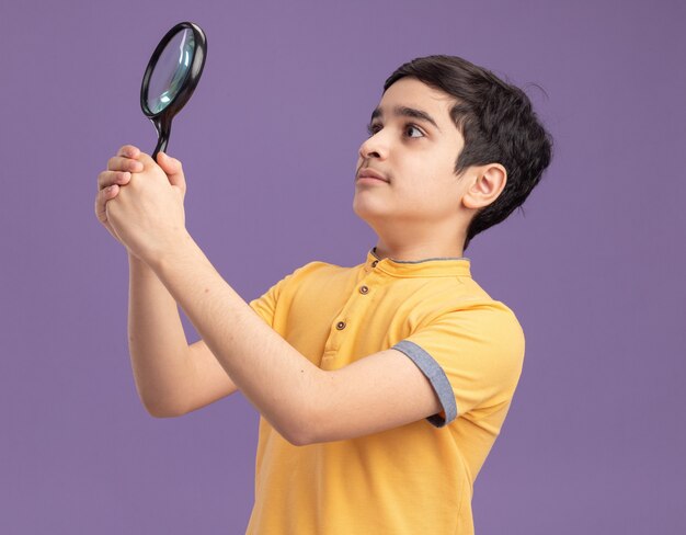 Jeune garçon caucasien impressionné tenant et regardant le côté à travers une loupe isolée sur un mur violet