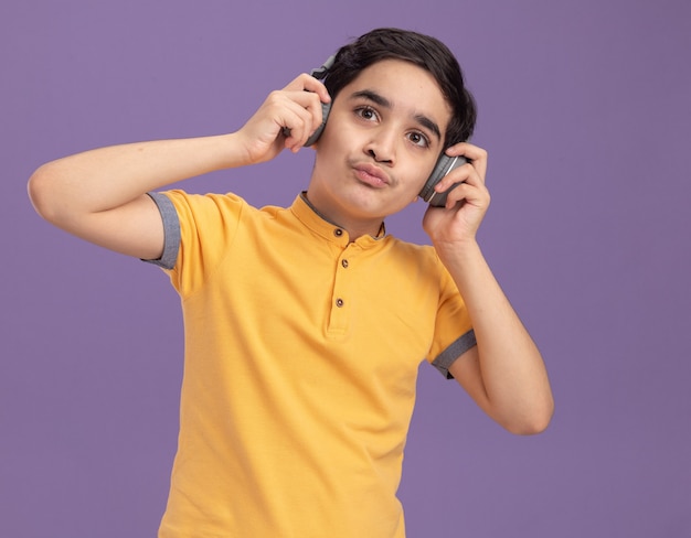 Jeune garçon caucasien portant et saisissant des écouteurs regardant droit en écoutant de la musique avec des lèvres pincées isolées sur un mur violet