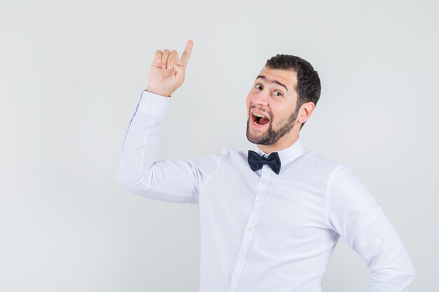 Jeune garçon en chemise blanche pointant le doigt vers le haut et à la vue de face, heureux.