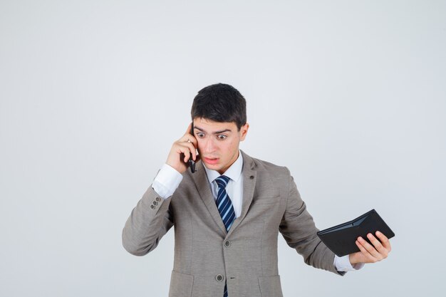 Jeune garçon en costume formel parlant au téléphone, tenant la calculatrice et à la surprise, vue de face.