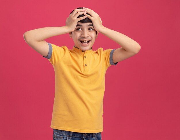 Jeune garçon impressionné mettant les mains sur la tête en regardant le côté isolé sur le mur rose