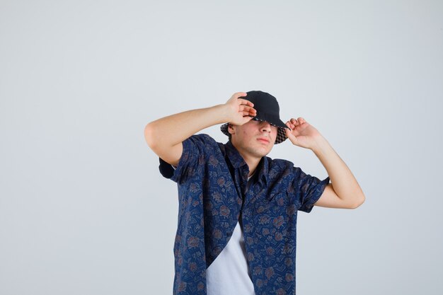 Jeune garçon mettant les mains sur la casquette en t-shirt blanc, chemise à fleurs, casquette et à la confiance. vue de face.