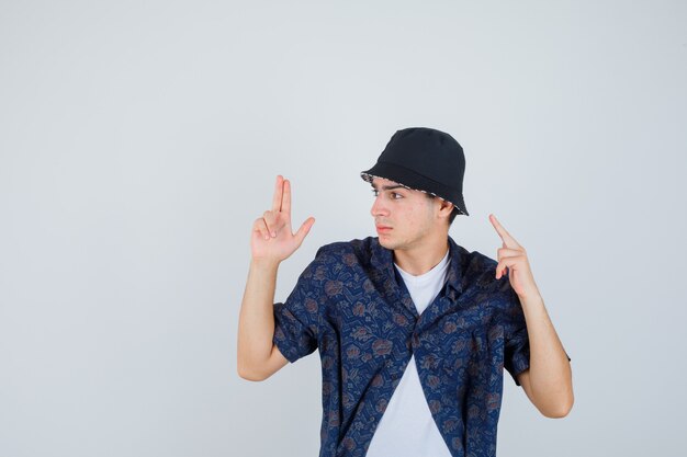 Jeune garçon montrant le geste du pistolet, levant l'index en t-shirt blanc, chemise fleurie, casquette et ayant l'air confiant.