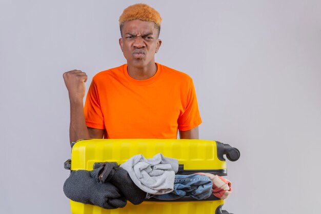 Jeune garçon portant un t-shirt orange debout avec une valise de voyage pleine de vêtements mécontents de froncer les sourcils serrant le poing sur le mur blanc