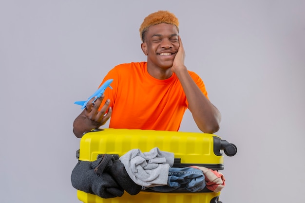 Jeune garçon portant un t-shirt orange debout avec une valise de voyage pleine de vêtements et tenant un avion jouet