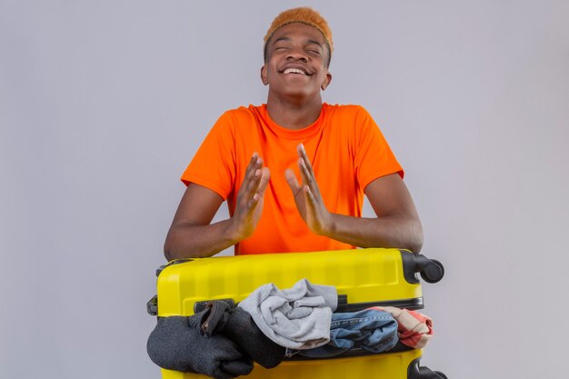 Jeune garçon portant un t-shirt orange debout avec les yeux fermés avec valise de voyage pleine de vêtements se tenant la main avec un visage heureux sur un mur blanc