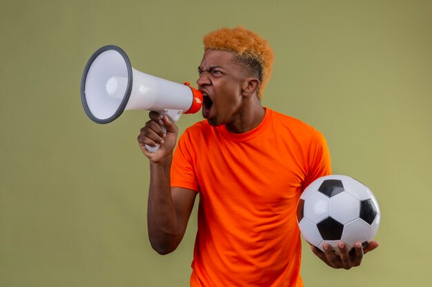 Photo gratuite jeune garçon portant un t-shirt orange tenant un ballon de football criant au mégaphone avec une expression de colère sur le visage debout sur un mur vert