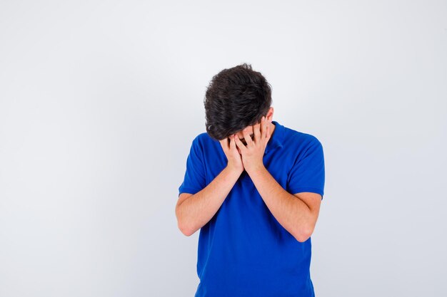 Jeune garçon en t-shirt bleu couvrant une partie du visage avec la main et l'air ennuyé, vue de face.