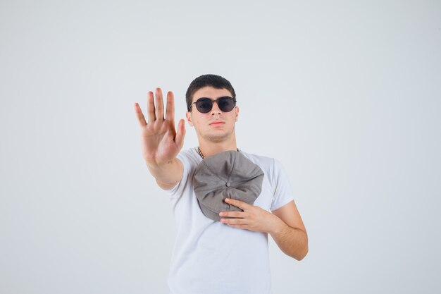 Jeune garçon tenant un chapeau sur la poitrine tout en montrant la paume en t-shirt et à la confiance en soi. vue de face.