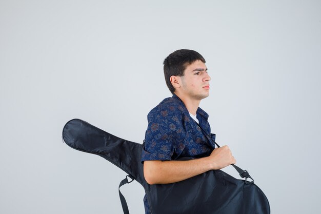 Jeune garçon tenant la guitare, regardant de côté en t-shirt et à la recherche concentrée. vue de face.