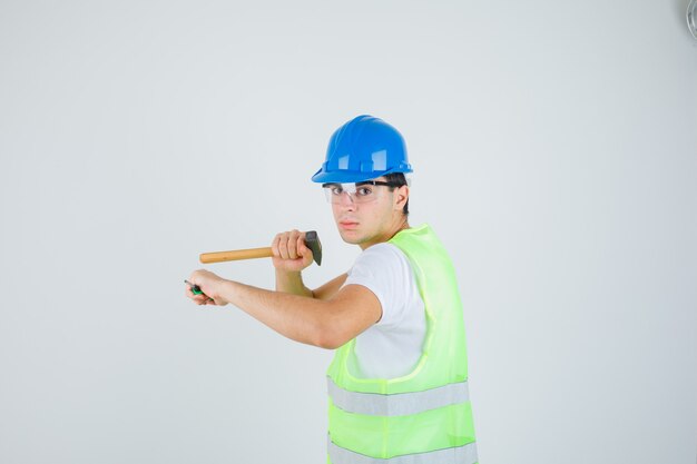Jeune garçon tenant un marteau et un tournevis en uniforme de construction et à la confiance. vue de face.