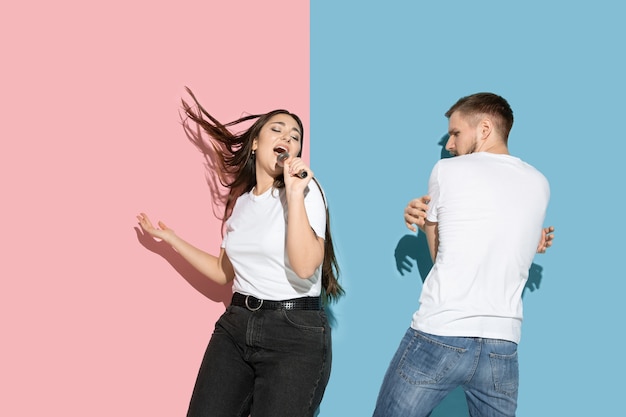 Jeune et heureux homme et femme dans des vêtements décontractés sur un mur bicolore rose, bleu, chant et danse