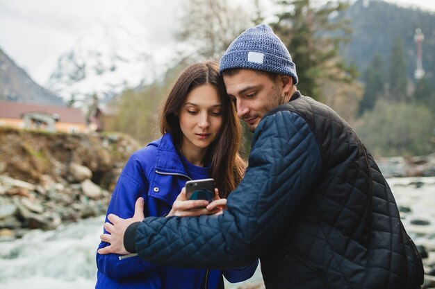 Jeune hipster beau couple amoureux tenant le smartphone, prendre des photos, à la rivière dans la forêt d'hiver