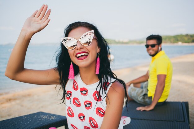 Jeune hipster élégant belle femme assise sur la plage, flirty, sexy, chaud, tenue de mode, lunettes de soleil à la mode, vacances tropicales, romance de vacances, homme sur fond à la recherche, souriant, agitant la main