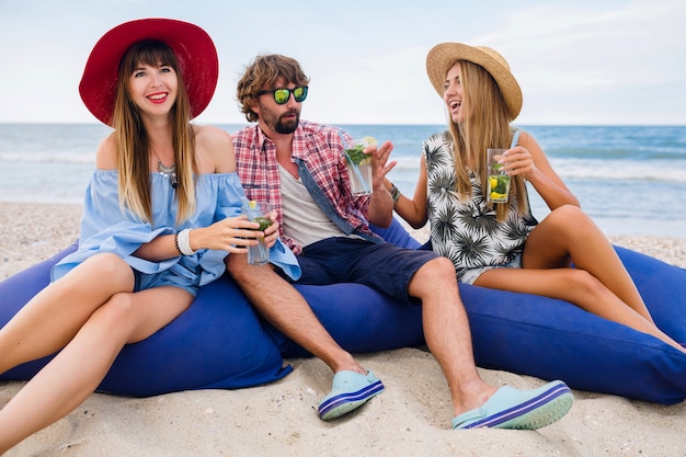 Jeune hipster souriant compagnie d'amis heureux en vacances assis dans des sacs de haricots sur une fête à la plage, boire un cocktail mojito
