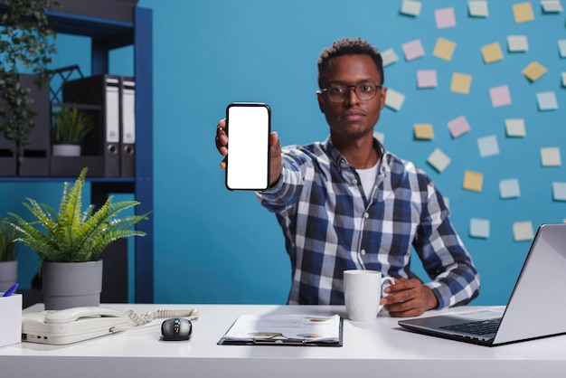 Jeune homme d'affaires adulte assis dans le bureau de l'espace de travail moderne de l'entreprise tout en montrant un modèle isolé d'écran blanc à la caméra. Chef de projet pointant un appareil téléphonique avec un écran de maquette vers l'appareil photo.