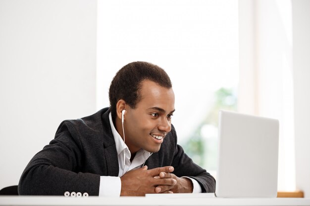 Jeune homme d'affaires africain réussi dans les écouteurs souriant, regardant un ordinateur portable.