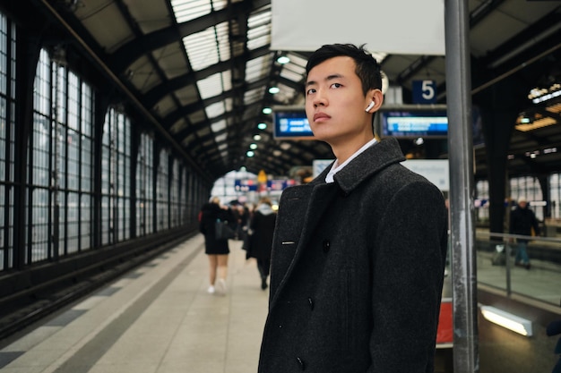 Photo gratuite jeune homme d'affaires asiatique élégant dans des écouteurs sans fil attendant le train à la station de métro