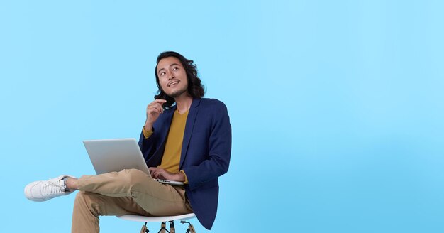 Jeune homme d'affaires asiatique pensant pendant qu'elle utilise un ordinateur portable assis sur une chaise sur fond bleu