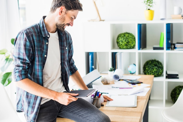 Jeune homme d&#39;affaires assis sur le bureau en tenant le bloc-notes