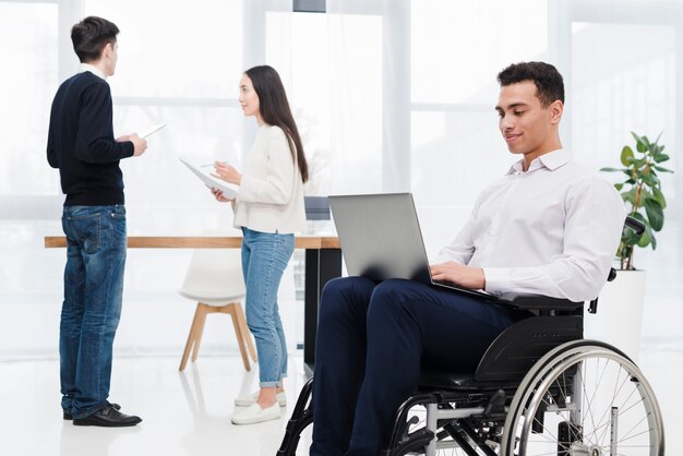 Jeune homme d&#39;affaires assis sur un fauteuil roulant à l&#39;aide d&#39;un ordinateur portable avec son collègue discutant de quelque chose à l&#39;arrière-plan