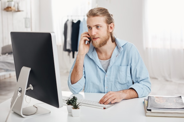 Un jeune homme d'affaires blond et barbu qualifié travaille sur un nouveau projet, s'assoit devant l'écran, a une conversation téléphonique et discute du rapport financier avec un partenaire commercial. Employé de bureau discute avec le patron