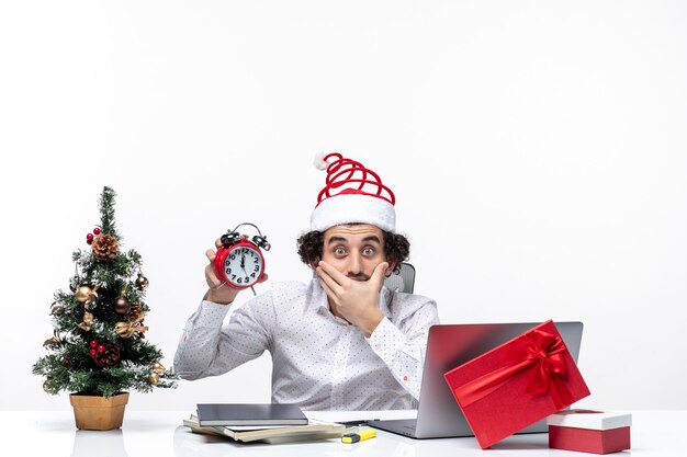 Jeune homme d'affaires avec chapeau de père Noël et montrant l'horloge en pensant à quelque chose avec soin et assis au bureau sur fond blanc