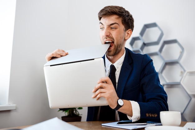 Jeune homme d'affaires en colère rongeant un ordinateur portable, fond de bureau.
