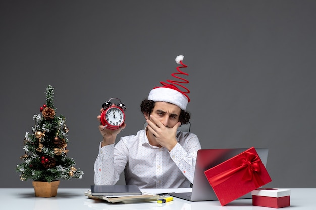 Jeune homme d'affaires concerné avec chapeau de père Noël et tenant une horloge et penser à quelque chose au bureau sur fond sombre