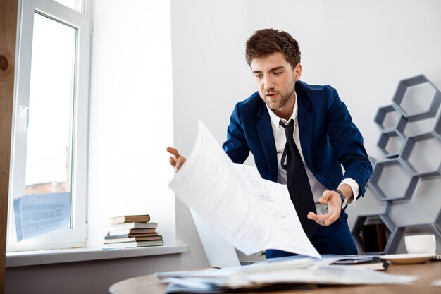 Jeune homme d'affaires distrait fouillant dans les papiers, fond de bureau.