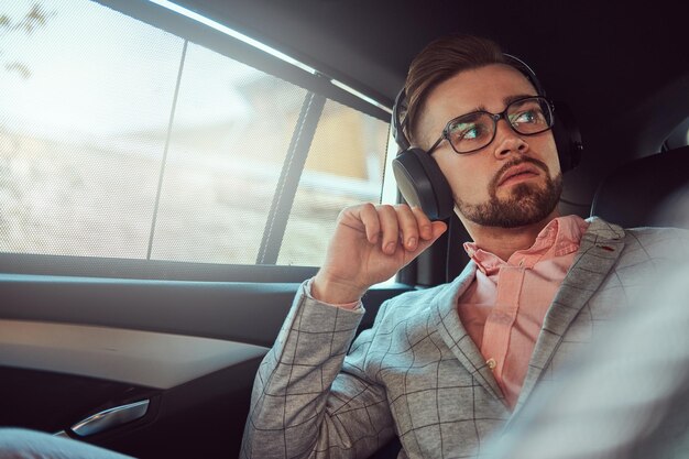 Jeune homme d'affaires élégant et prospère dans un costume gris et une chemise rose, écoutant de la musique sur des écouteurs, monté sur un siège arrière dans une voiture de luxe.