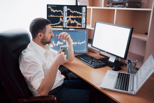 Jeune homme d'affaires est assis au bureau à table, travaillant sur ordinateur avec de nombreux moniteurs, diagrammes sur moniteur. Courtier en bourse analyse les graphiques d'options binaires.Hipster homme buvant du café, étudiant