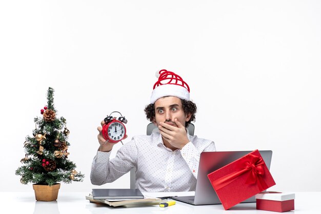 Jeune homme d'affaires excité avec chapeau de père Noël et montrant l'horloge travaillant seul assis au bureau sur fond blanc