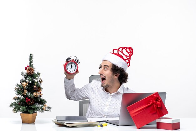 Jeune homme d'affaires excité émotionnel drôle avec chapeau de père Noël et tenant et montrant l'horloge assis au bureau sur fond blanc