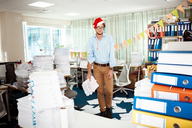 Jeune homme d'affaires fatigué travaillant au bureau parmi les papiers le jour de Noël.