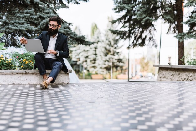 Jeune homme d&#39;affaires moderne assis dans le parc à la recherche d&#39;un ordinateur portable