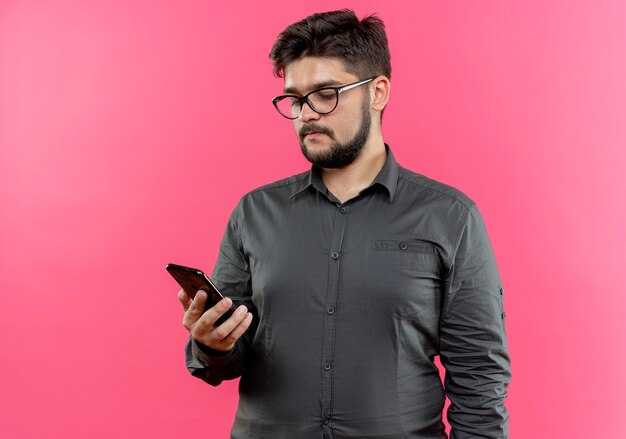 Jeune homme d'affaires portant des lunettes tenant et regardant le téléphone isolé sur rose