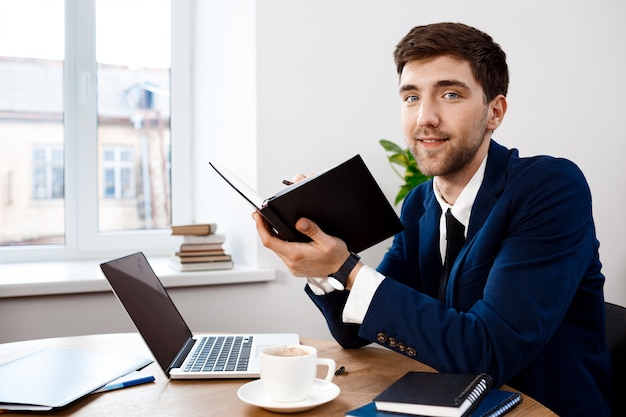 Jeune homme d'affaires prospère, assis sur le lieu de travail, fond de bureau.