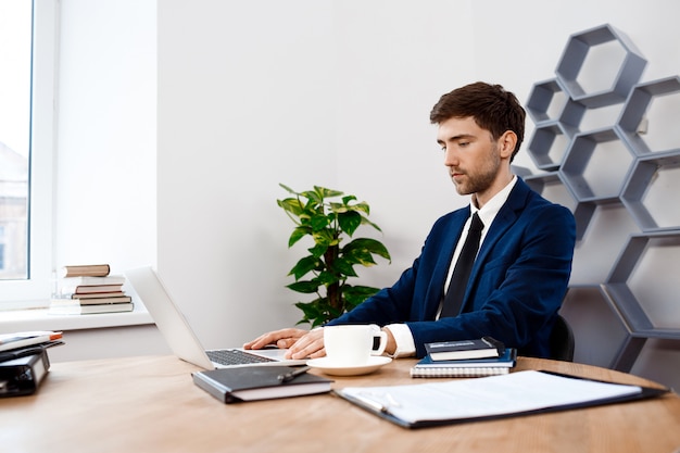 Jeune homme d'affaires prospère, assis sur un ordinateur portable, fond de bureau.