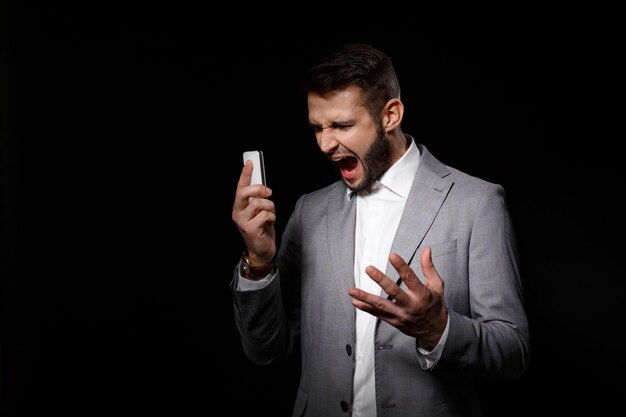 Jeune homme d'affaires prospère en costume criant en regardant le téléphone sur fond noir