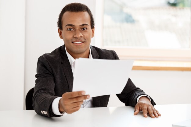 Jeune homme d'affaires prospère souriant, tenant du papier, assis sur le lieu de travail