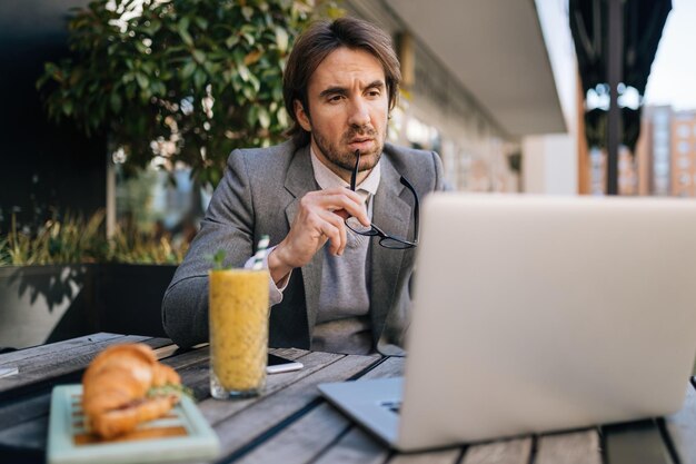 Jeune homme d'affaires réfléchi travaillant sur un ordinateur assis dans un café en plein air