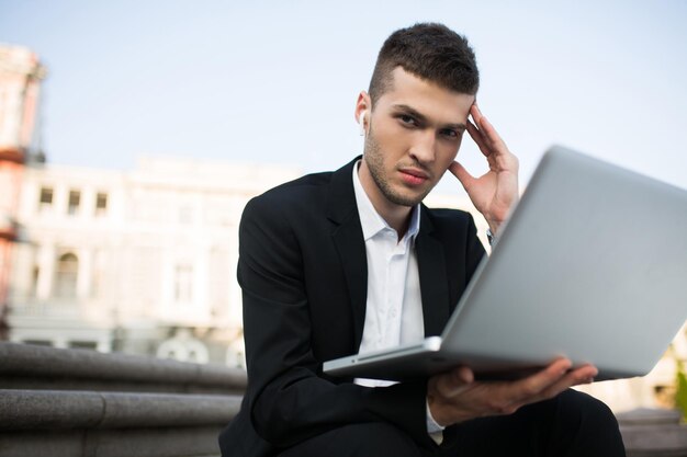 Jeune homme d'affaires sérieux en veste noire classique et chemise blanche avec des écouteurs sans fil regardant pensivement à huis clos avec un ordinateur portable à la main en plein air