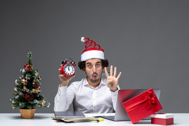 Jeune homme d'affaires surpris concerné avec chapeau de père Noël et tenant une horloge et montrant quatre dans le bureau sur fond sombre