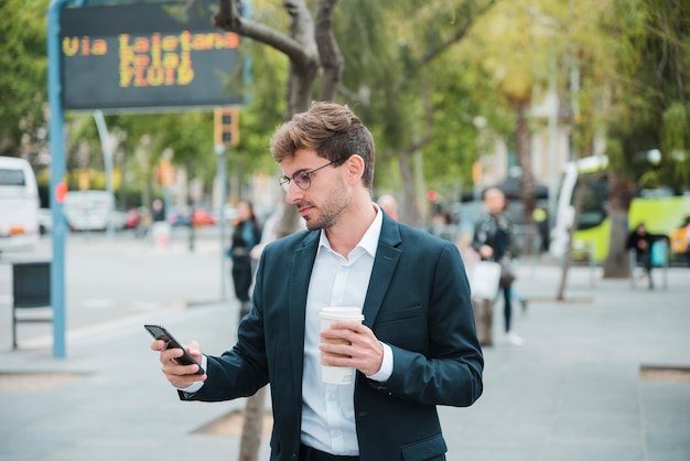 Jeune homme d&#39;affaires tenant une tasse de café jetable en regardant téléphone mobile