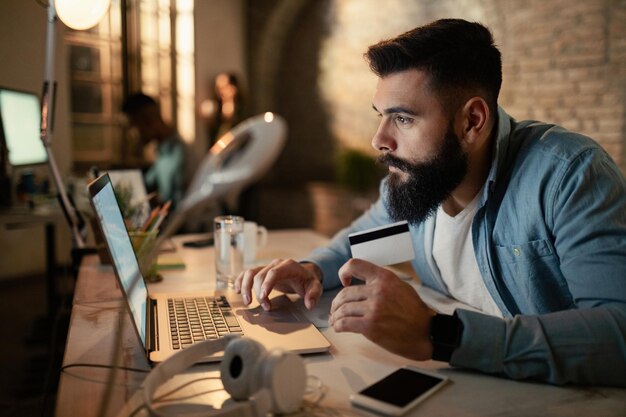 Jeune homme d'affaires utilisant un ordinateur portable et une carte de crédit tout en achetant sur Internet la nuit au bureau