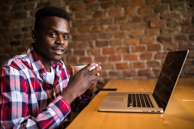 Photo gratuite jeune homme africain assis au café avec ordinateur portable. étudiant portant des vêtements à la mode, buvant du café, utilisant internet sans fil. concept de technologie et de communication
