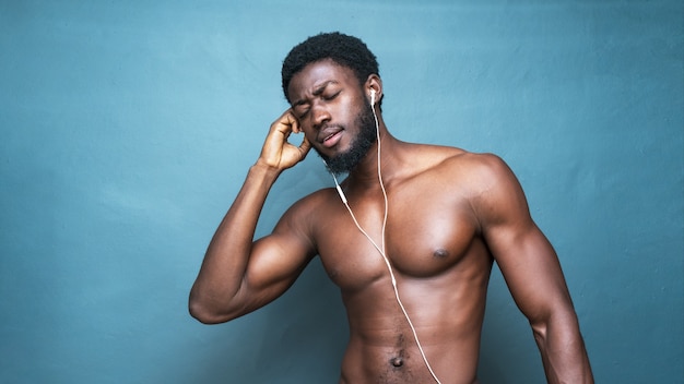 Jeune homme africain aux seins nus chaud écoutant de la musique avec des écouteurs sur un bleu